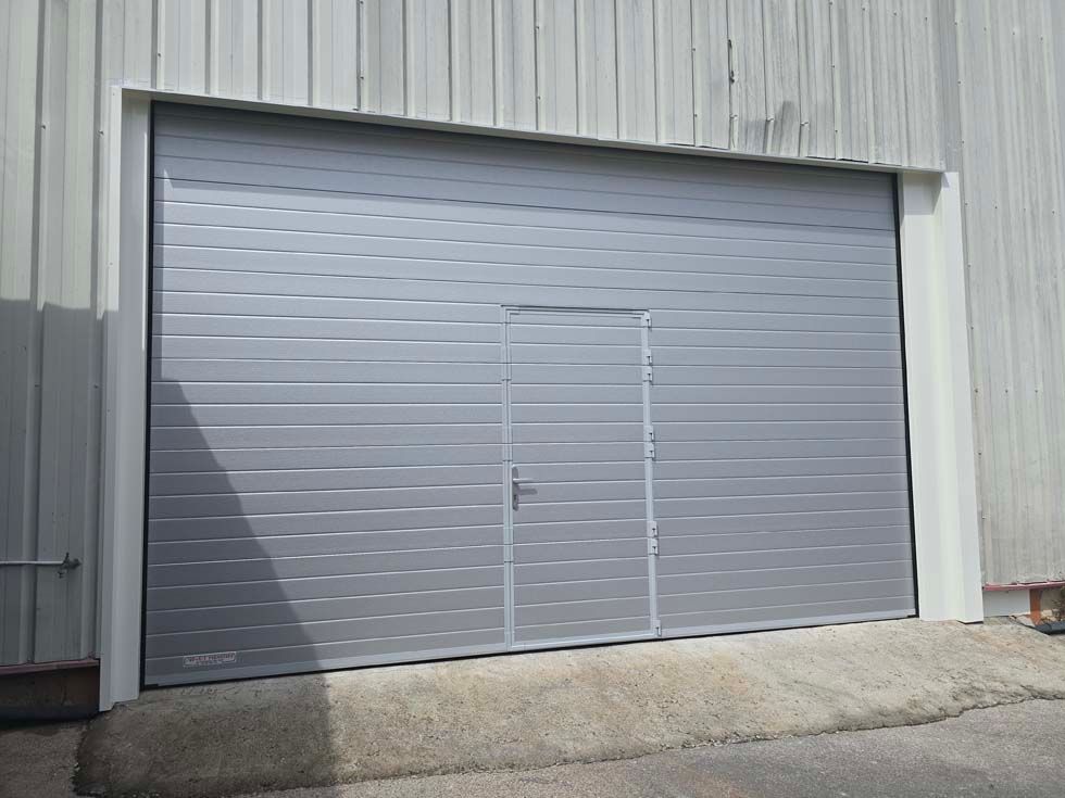 Grande porte de garage industrielle grise avec une porte intégrée plus petite, installée dans une ouverture à cadre blanc.