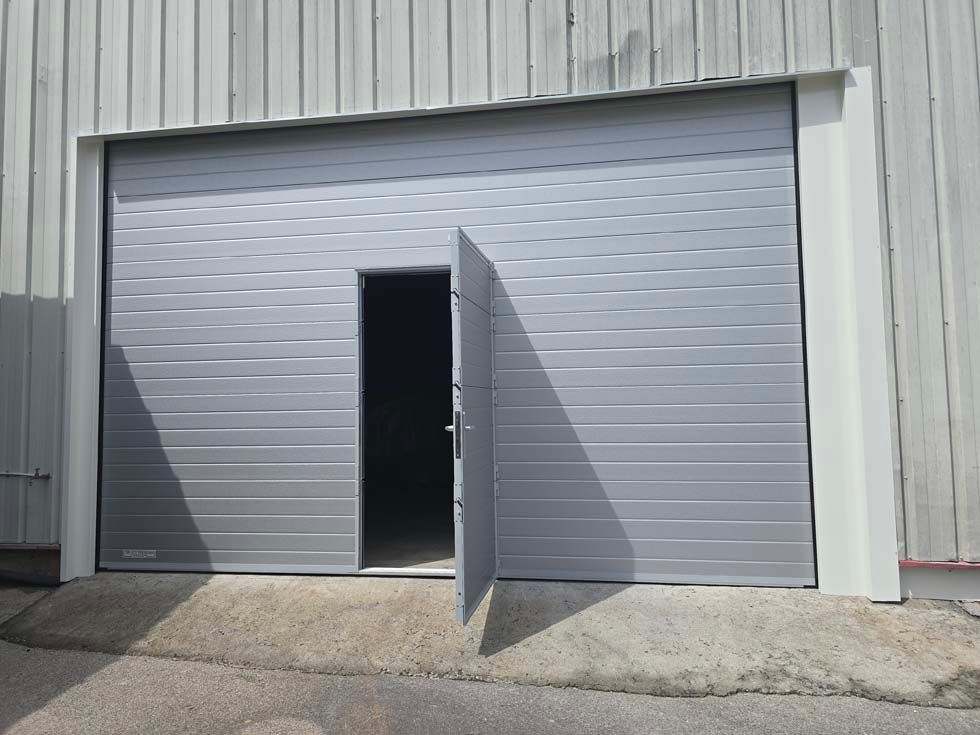 Porte sectionnelle métallique grise avec une petite porte piétonne intégrée ; extérieur du bâtiment.