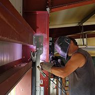 Un soudeur dans un atelier utilise une torche de soudage sur des poutres métalliques rouges.
