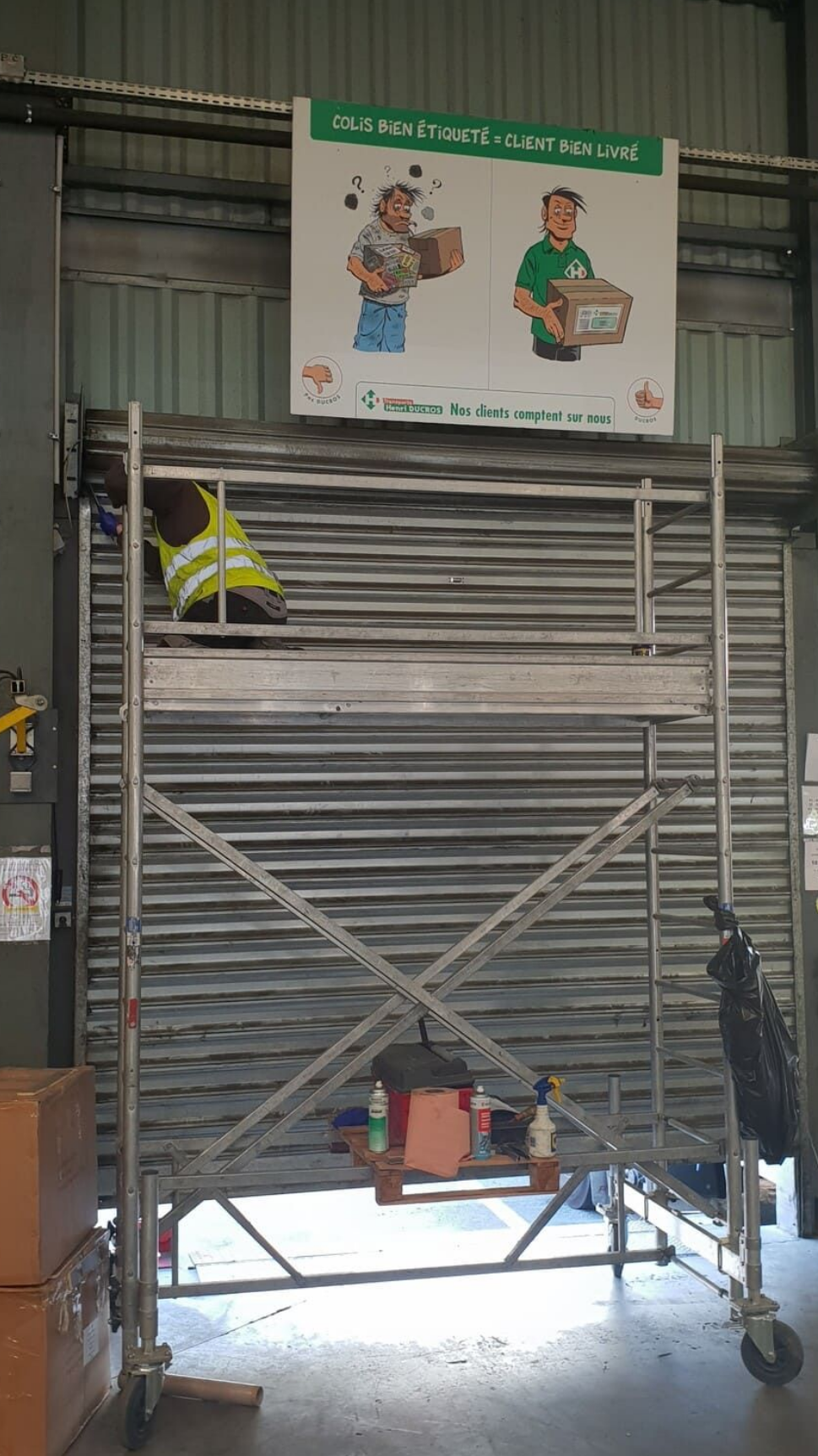 Échafaudage dans un entrepôt avec panneau de sécurité indiquant les techniques de levage appropriées. Ouvrier en gilet jaune.