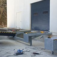 Quai de chargement avec porte sectionnelle grise partiellement ouverte, rampe métallique et outils au sol.