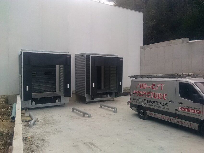 Deux grands conteneurs industriels devant un bâtiment blanc, à côté d'une camionnette.