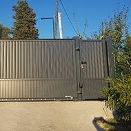 Portail fermé en métal gris foncé à rayures verticales, sur une allée en béton.