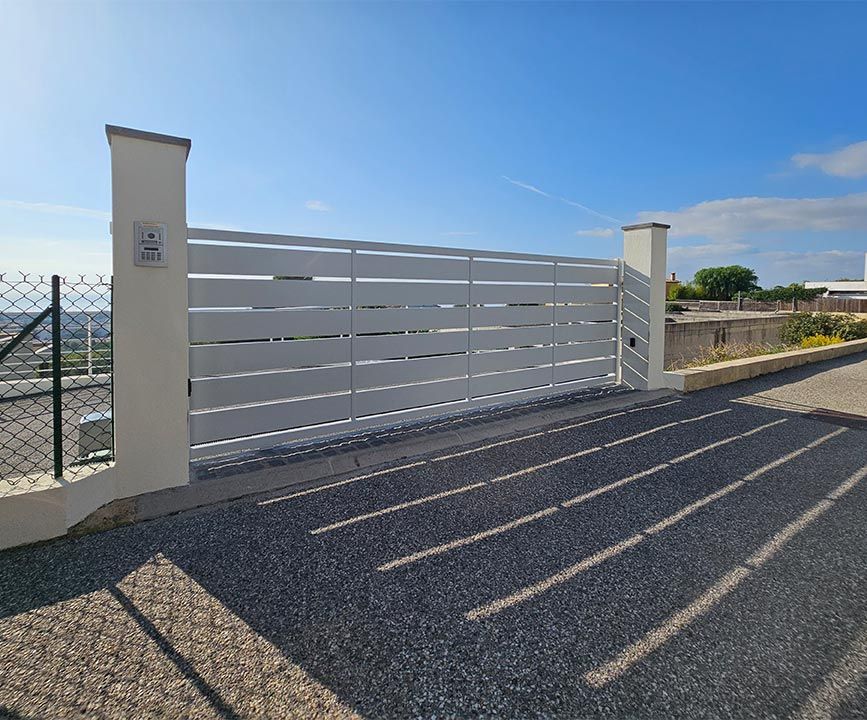 Portail à lattes horizontales blanches entre deux piliers blancs sur une allée de gravier.