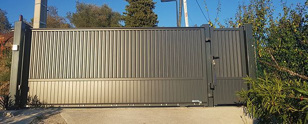 Portail métallique dans un cadre ensoleillé. Le portail est fermé et présente des lignes verticales et ondulées. On aperçoit des buissons et le ciel.