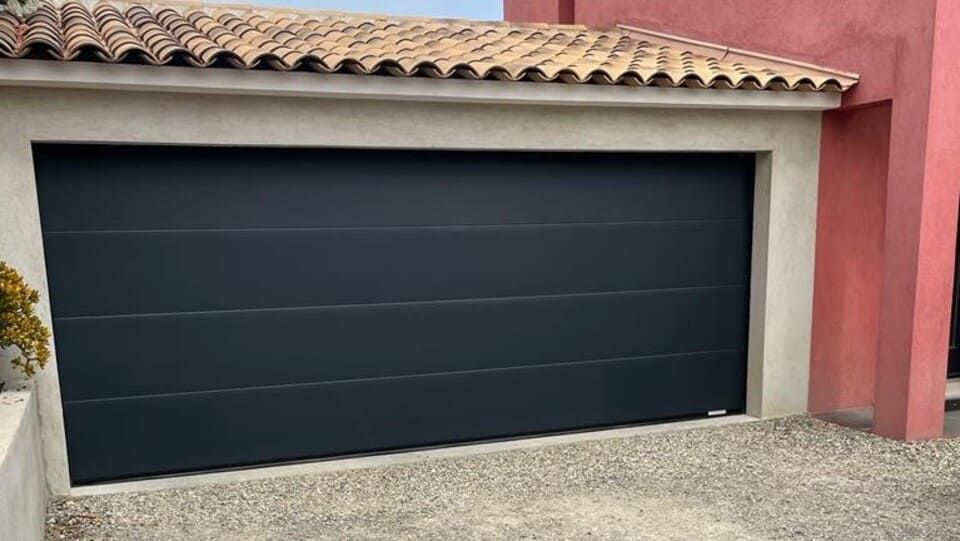 Porte de garage gris foncé sur une maison avec un toit de tuiles et une allée en gravier.
