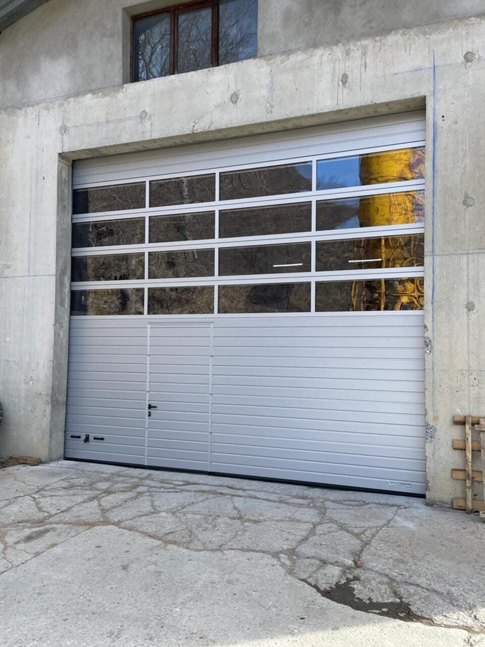 Porte de garage avec partie supérieure transparente, panneaux argentés, dans un bâtiment en béton.