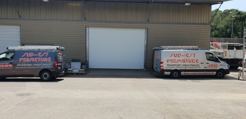 Deux camionnettes sont garées devant un entrepôt. L'une est grise avec des inscriptions rouges, l'autre est blanche avec des inscriptions rouges.