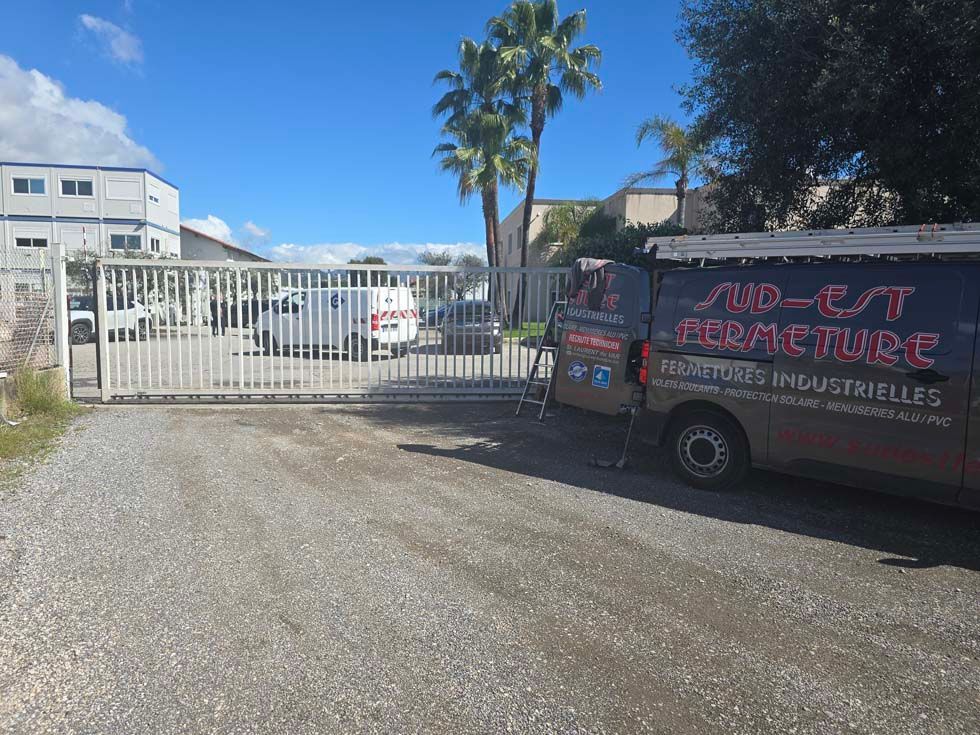 Allée de gravier avec portail métallique ouvert ; camionnette blanche garée à droite ; ciel bleu et palmiers en arrière-plan.