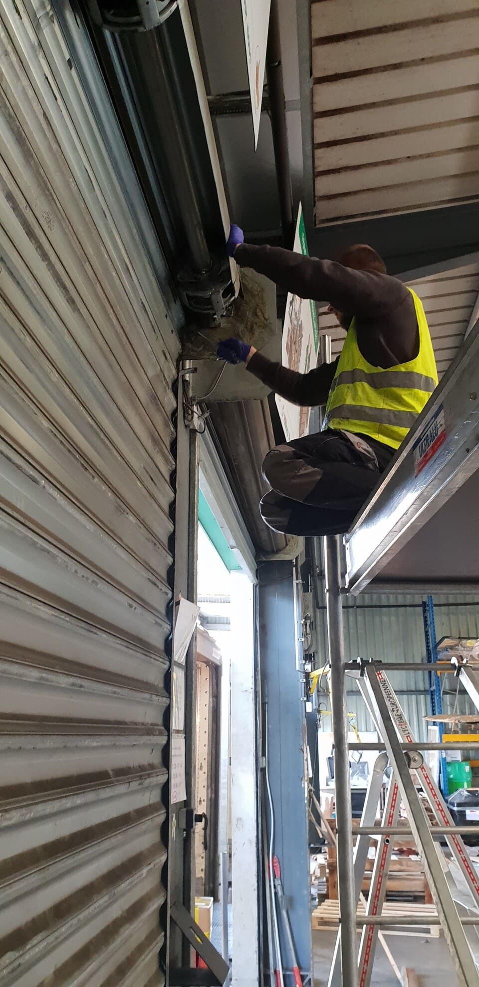 Une personne portant un gilet de sécurité travaille sur une porte roulante, à l'aide d'une échelle à l'intérieur d'un bâtiment.