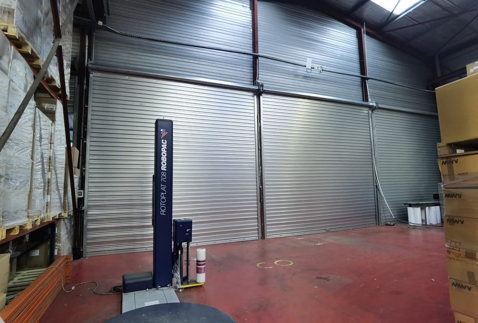 Intérieur d'un entrepôt avec une grande porte métallique, une banderoleuse de palettes et des cartons sur la droite.