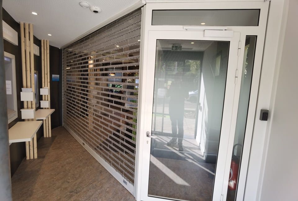 Hall d'entrée avec portail de sécurité partiellement ouvert, porte vitrée et personne visible.