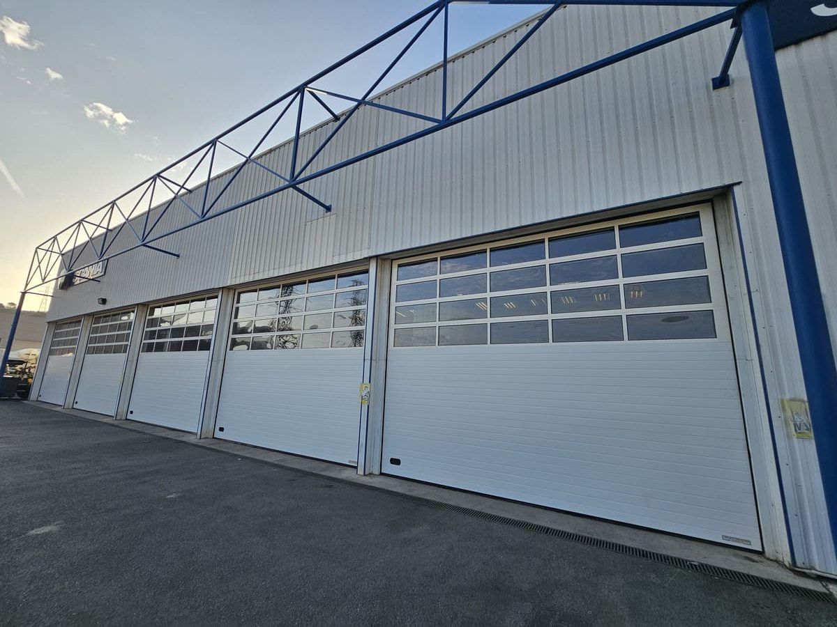 Extérieur d'un bâtiment industriel léger avec de grandes portes de garage blanches et une structure en acier bleu au-dessus.