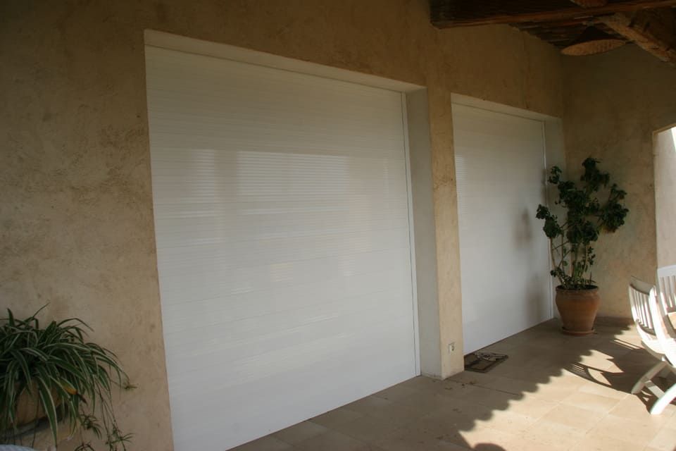 Deux grands stores blancs enroulables sur un mur extérieur en stuc, à côté de plantes et d'une terrasse.