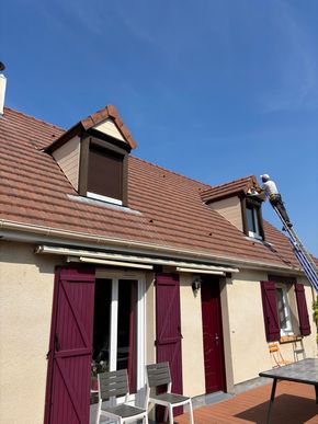 Un ouvrier, juché sur une échelle, répare le toit d'une maison rose aux volets rouges sous un ciel bleu.