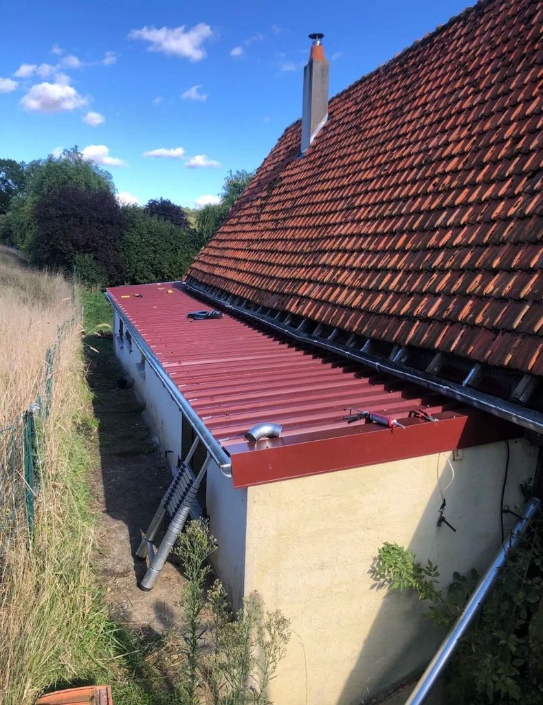 Maison avec toit de tuiles rouges et petite extension au toit rouge donnant sur un champ herbeux sous un ciel bleu