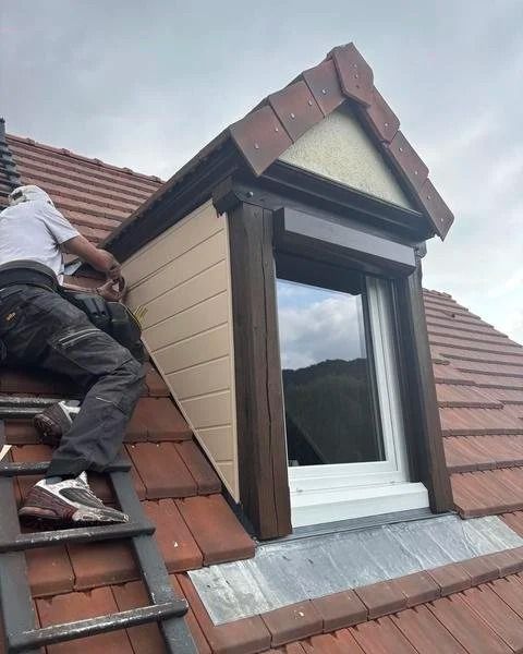 Ouvrier installant une lucarne sur un toit de tuiles, avec des moulures brunes et un cadre de fenêtre blanc