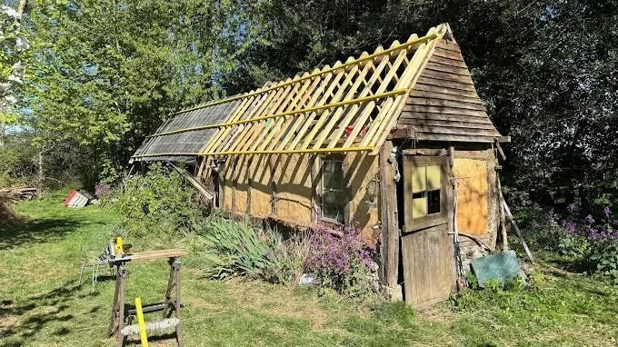 Petit abri rustique en bois en construction dans un jardin herbeux, avec la charpente apparente et des outils à proximité.