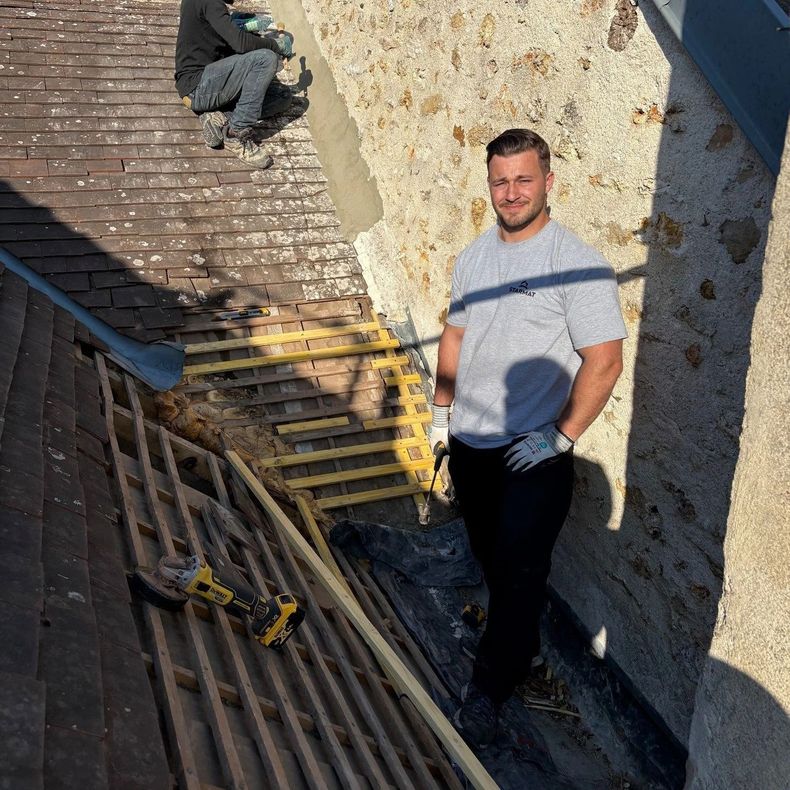 Un ouvrier se tient debout sur un toit en pente, près d'un mur, tandis qu'un autre artisan est accroupi près du bord.