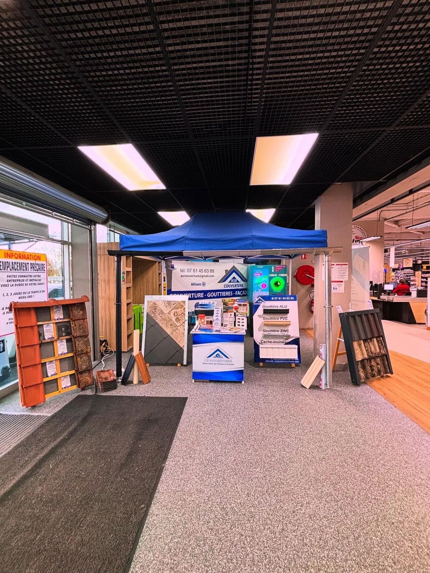 Stand d'exposition intérieur avec auvent bleu, affiches, panneaux et présentoirs à brochures dans un couloir.