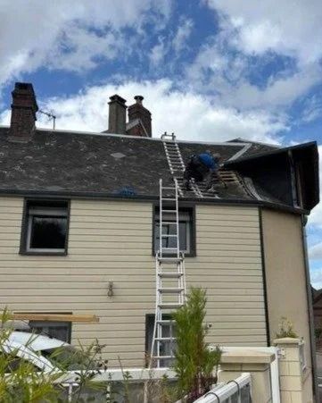 Un ouvrier, perché sur une échelle, répare le toit d'une maison beige sous un ciel nuageux.
