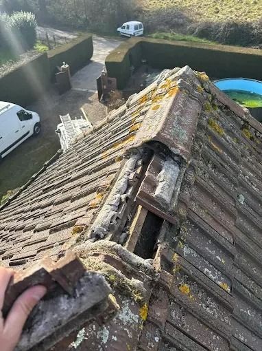 Toiture en tuiles endommagée, avec des tuiles faîtières manquantes, vue depuis le toit, donnant sur une allée et des camionnettes stationnées.