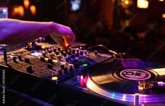 La main d'un DJ ajuste une table de mixage à côté d'une platine incandescente dans un club à l'ambiance tamisée.