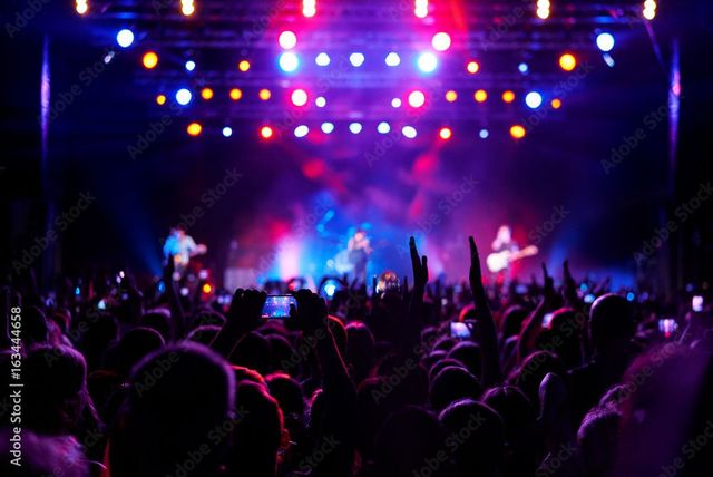 Foule à un concert nocturne, les mains levées, sous les lumières colorées de la scène qui brillent en violet, bleu et rose.