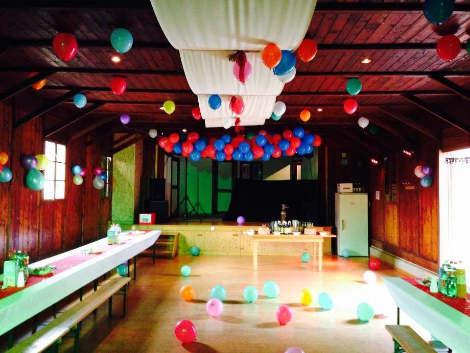 Salle remplie de ballons avec de longues tables, des décorations suspendues colorées et une petite scène au fond.