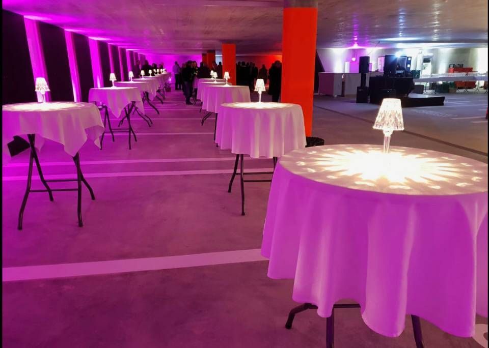 Salle de réception vide avec des tables rondes drapées de guirlandes lumineuses blanches et violettes, éclairage rose indirect le long des murs.