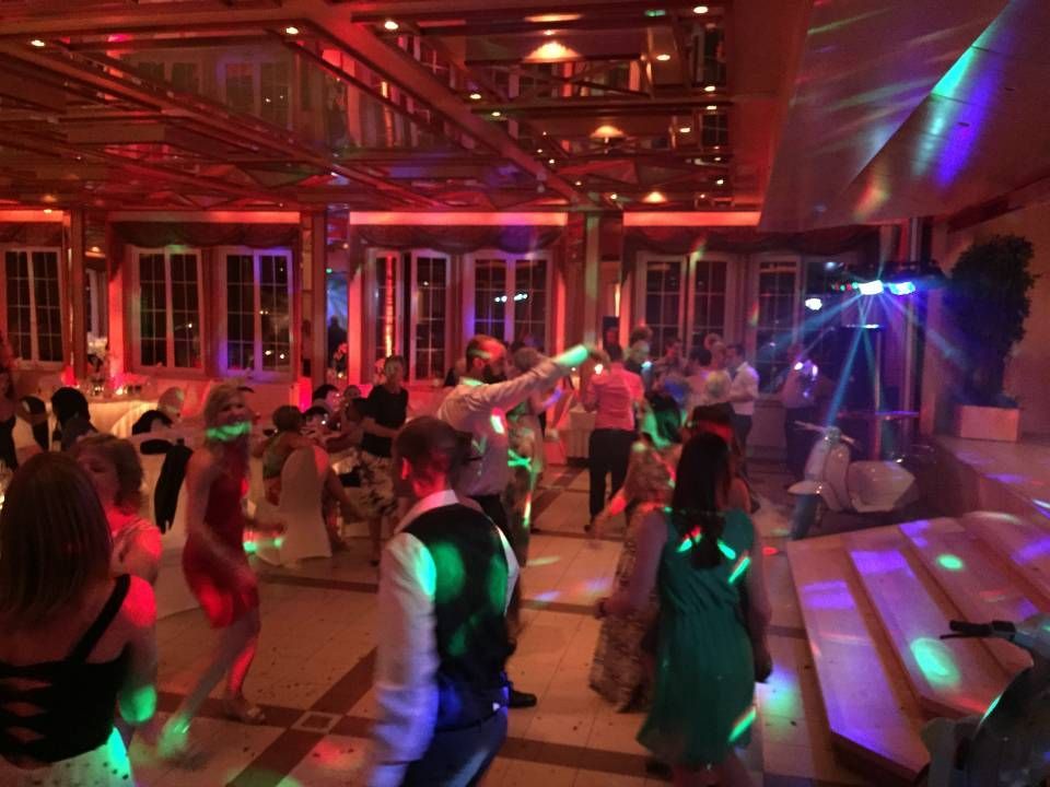 Des personnes dansent dans une salle de banquet éclairée en rouge, avec des lumières disco colorées et des marches d'estrade.