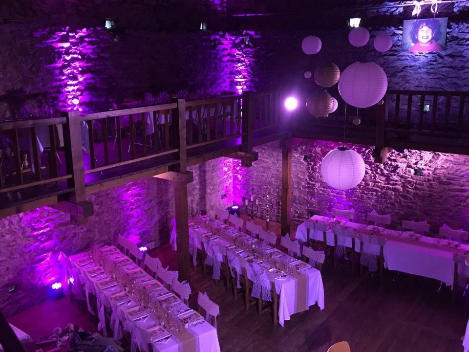 Salle de banquet éclairée de violet, avec de longues tables dressées pour un événement sous des murs de pierre et des balustrades de balcon.