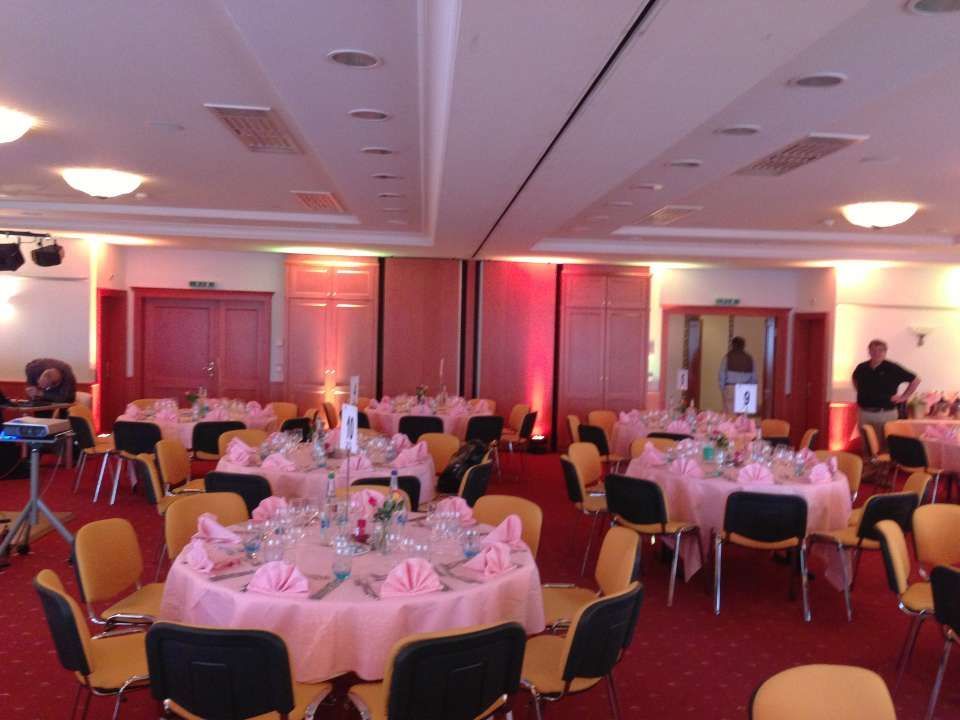 Salle de banquet ronde avec nappes roses et chaises dressées pour un événement, avec quelques personnes en arrière-plan.