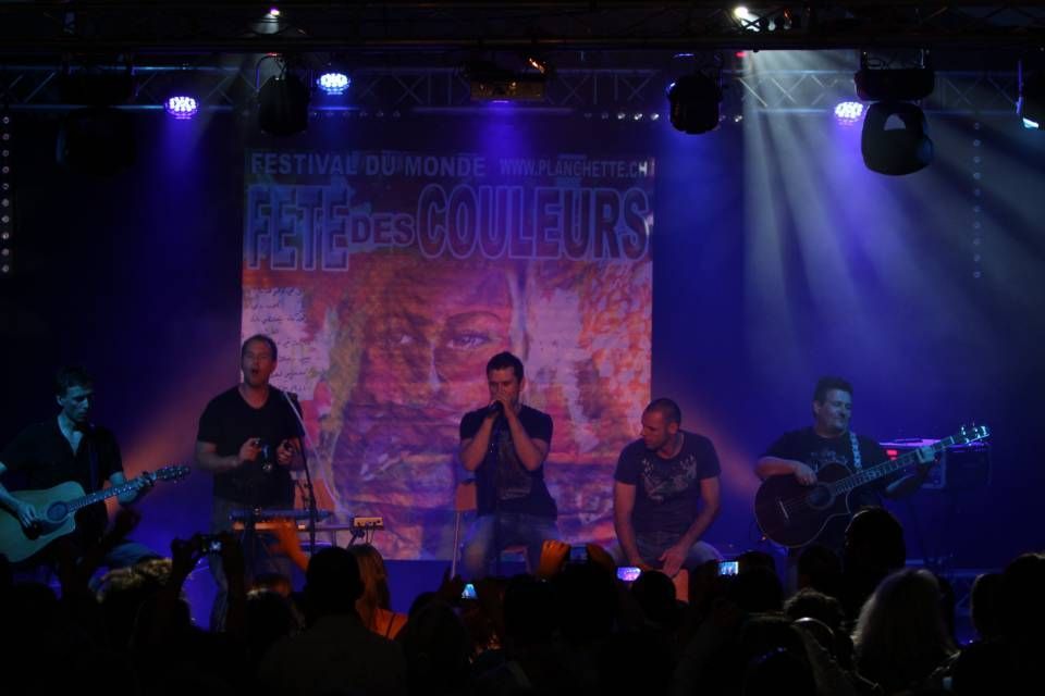 Un groupe de musique se produit en direct sur une scène faiblement éclairée de lumières bleues et violettes, le public se détachant en silhouette au premier plan.