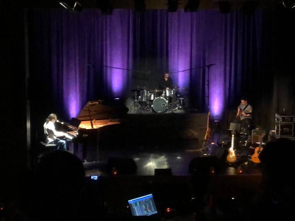 Un groupe de musique en live sur une scène sombre éclairée de violet, la batterie au centre, le piano à gauche et les guitares à droite.