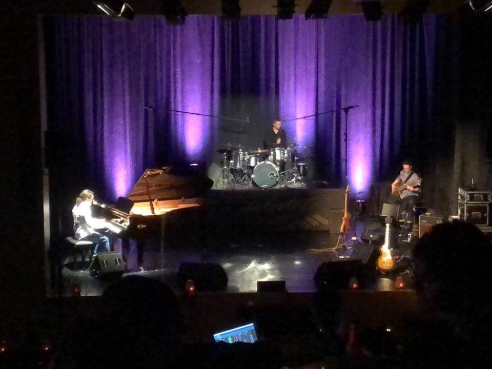 Scène de concert avec pianiste, batteur et bassiste sous une lumière violette