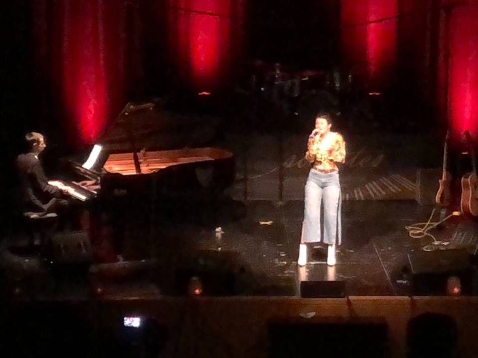 Une chanteuse sur une scène obscure éclairée de lumières rouges, debout à côté d'un pianiste à un piano à queue.