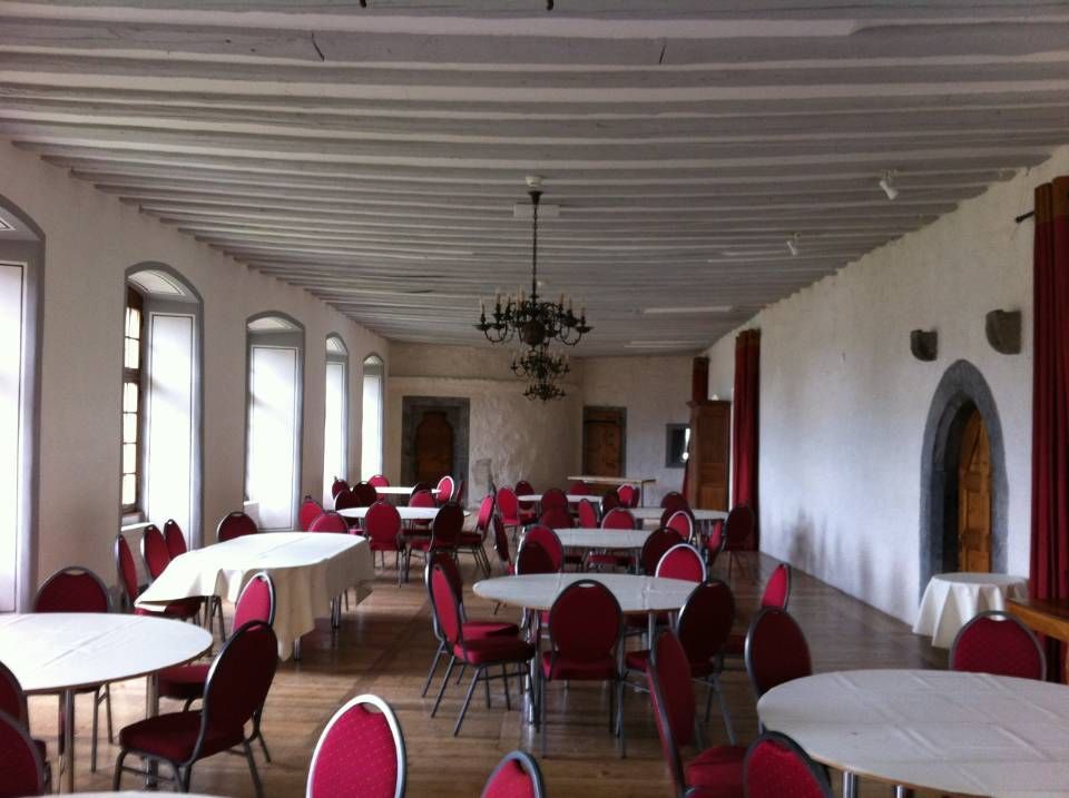 Salle à manger vide avec chaises rouges, nappes blanches, fenêtres cintrées et un lustre.