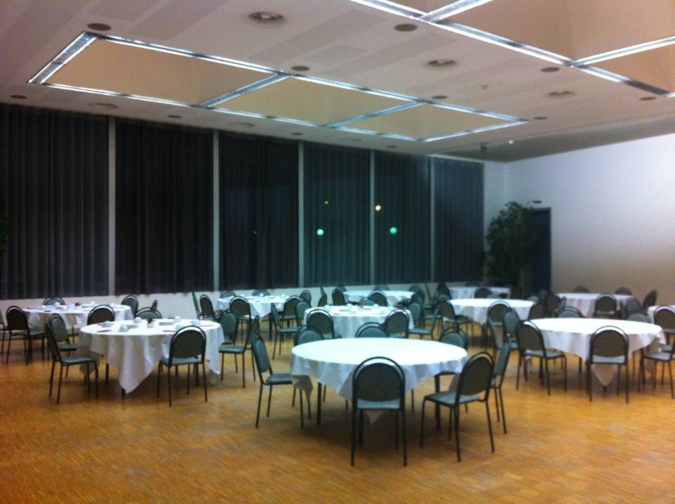 Salle de banquet vide avec tables rondes, nappes blanches, chaises noires et parquet sous des spots encastrés au plafond