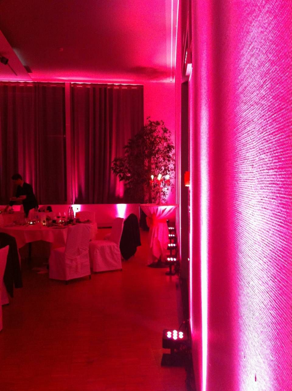 Salle de banquet éclairée de rose avec des tables rondes, des rideaux rouges et un arbre décoratif près du mur du fond.