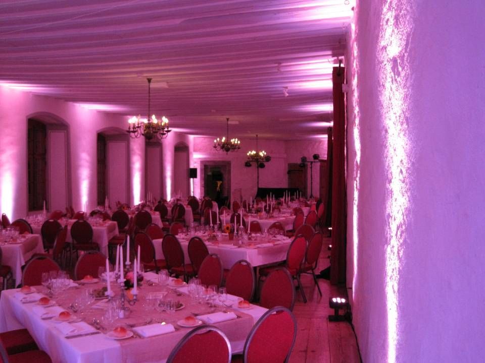 Salle de banquet éclairée de rose, avec tables rondes, chaises rouges et lustres, le long d'une longue pièce ornée de colonnes.