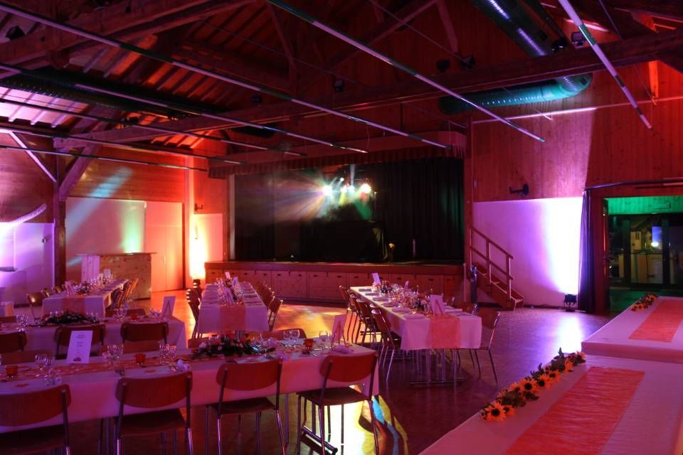 Salle de banquet vide avec tables et chaises blanches, éclairée par des projecteurs de scène rouges et violets