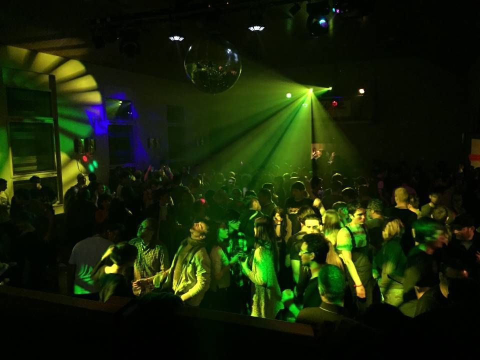 Piste de danse bondée d'une boîte de nuit, illuminée par des projecteurs verts et jaunes et surmontée d'une boule disco.