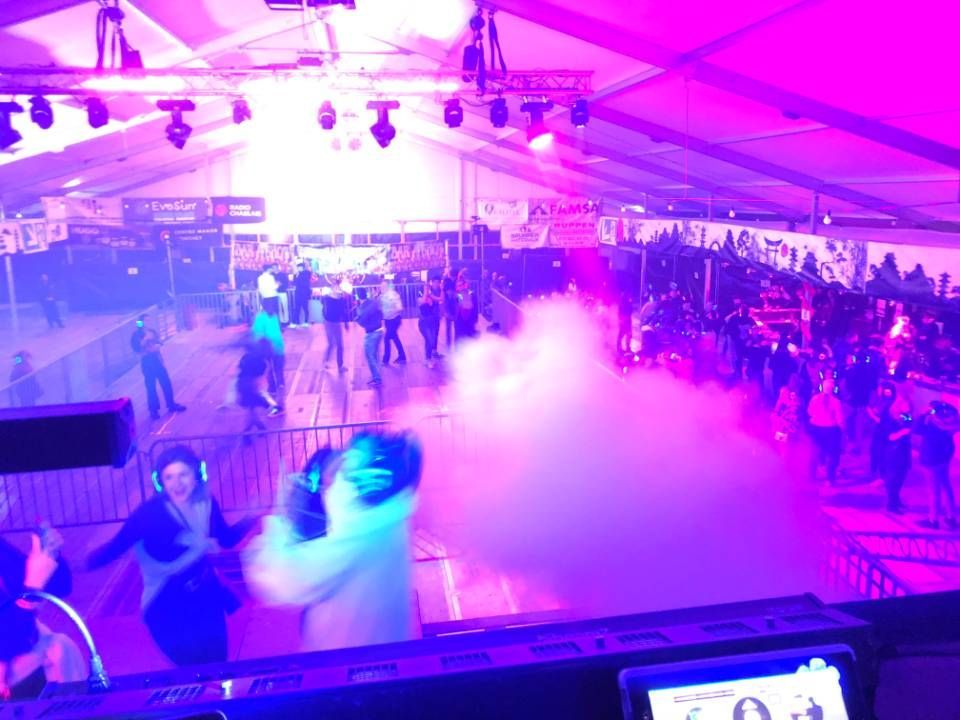 Des gens dansent sous des lumières violettes dans une salle intérieure bondée, avec du brouillard et une cabine de DJ.