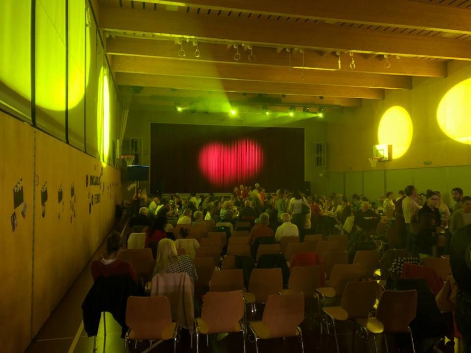 Le public est assis dans une salle faiblement éclairée, face à une scène ornée d'un rideau rouge et d'un éclairage jaune.