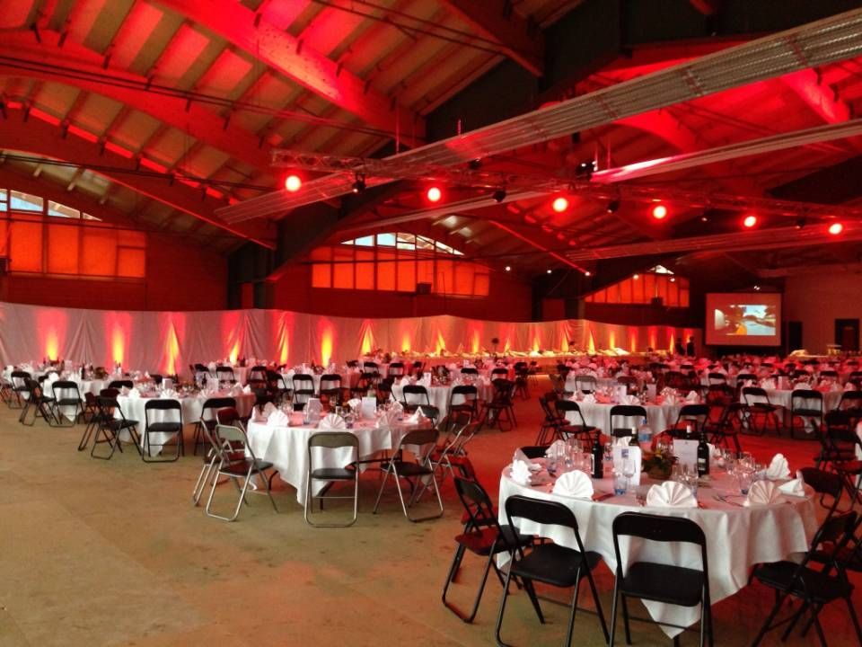 Salle de banquet éclairée en rouge, avec tables rondes et housses de chaises blanches, aménagée pour un grand événement.
