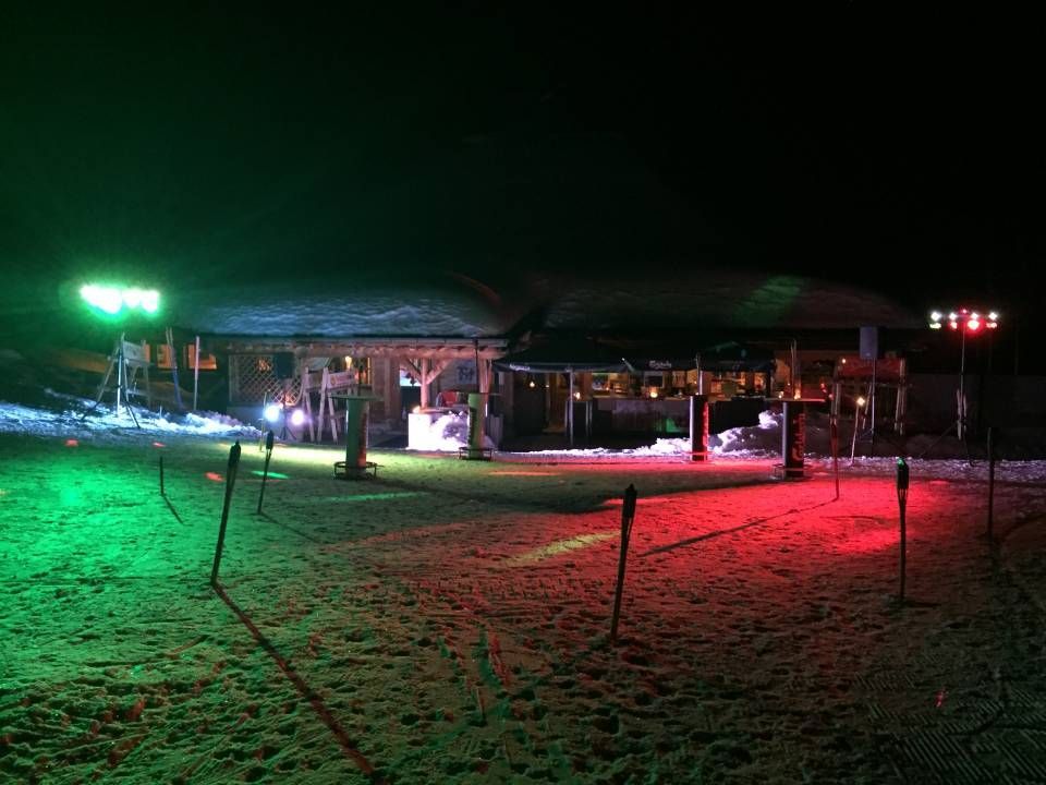 Scène nocturne en extérieur avec des lumières rouges et vertes illuminant une cour et un pavillon enneigés.