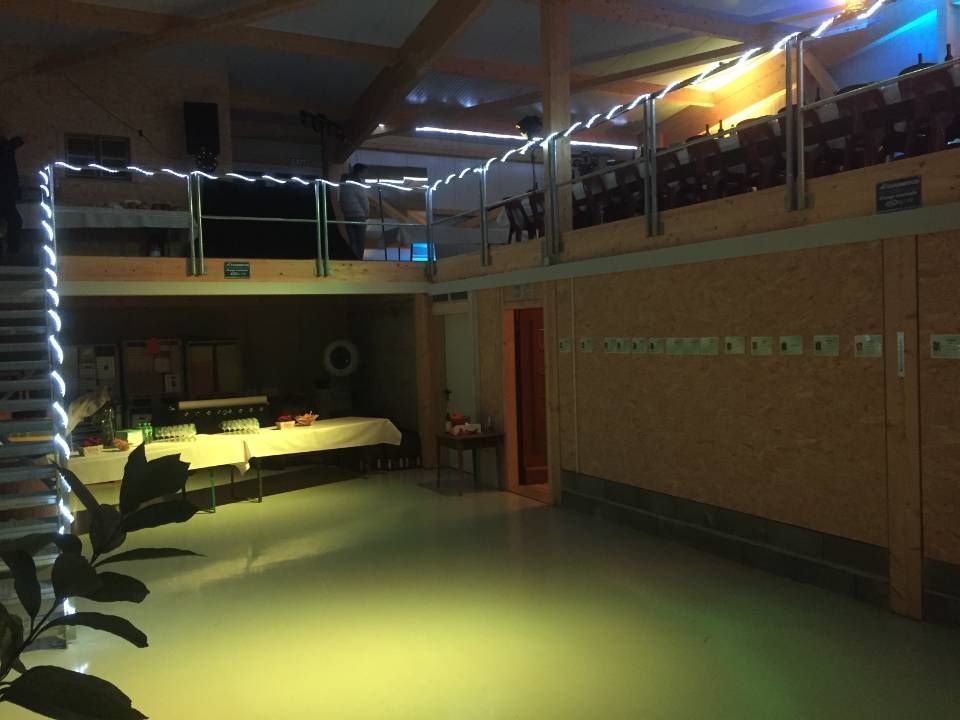 Hall intérieur tamisé avec éclairage chaleureux, guirlandes lumineuses, balcon en mezzanine et tables le long du mur de gauche.