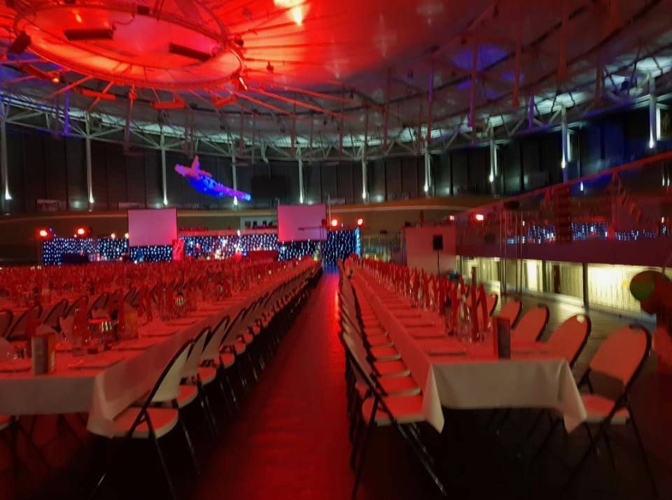 Salle de banquet éclairée en rouge, avec des rangées de tables et de chaises faisant face à une scène, vide et prête pour un événement.