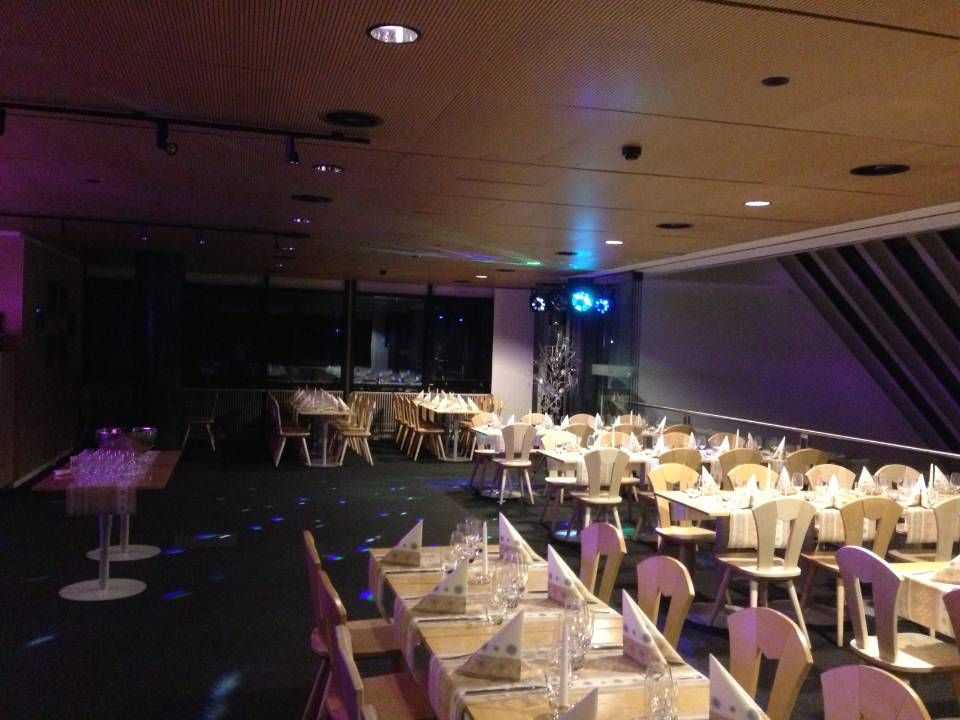 Salle de banquet faiblement éclairée, avec des rangées de tables et de chaises dressées, des murs violets et une lumière disco au fond.